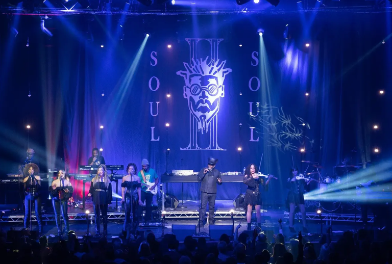 British R&B group Soul II Soul performing live on a concert stage under blue spotlights, backed by a large illuminated band logo backdrop.